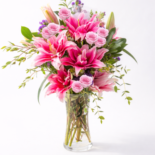 The Gazer Elegance from The Florist at Plantation features vibrant pink lilies, light pink roses, and assorted greenery arranged in a clear glass vase, set against a plain white background.