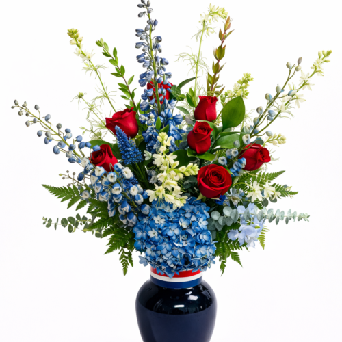 The American Honor Vase from The Florist at Plantation is a tall arrangement with red roses, blue delphiniums, white flowers, and lush greens in a dark blue vase—a striking Clayton NC Florist creation set against a white background.