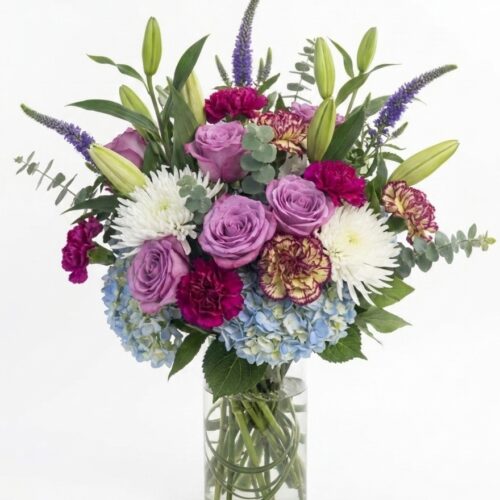 Blushing Meadows: A clear glass vase with a vibrant mix of purple roses, red and yellow carnations, white chrysanthemums, blue hydrangeas, green leaves, and purple veronica flowers on a white background.