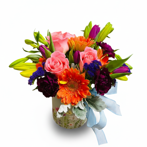 The Colorburst Gerbera Vase features vibrant pink roses, yellow lilies, orange gerberas, purple carnations, and lush greenery in a patterned vase with a light blue ribbon—perfect from your trusted Clayton NC Florist.