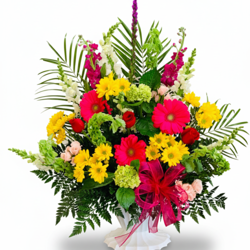 Remembrance in Bloom by The Florist at Plantation is a vibrant floral arrangement in a white vase with yellow daisies, pink gerberas, red roses, filler flowers, assorted greenery, and a large pink ribbon bow against a white background.
