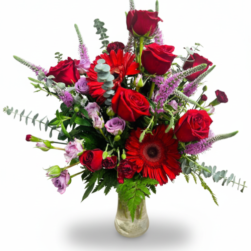 Red Petal Parade features a clear glass vase filled with red roses, gerbera daisies, purple and lavender blooms, and lush greenery, all expertly arranged by The Florist at Plantation against a white background.