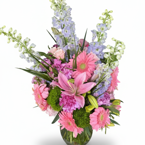 The Cotton Candy Garden Vase from The Florist at Plantation showcases pink gerbera daisies, lilies, carnations, light purple delphiniums, green pom-poms, and assorted greenery in a glass vase against a white background.