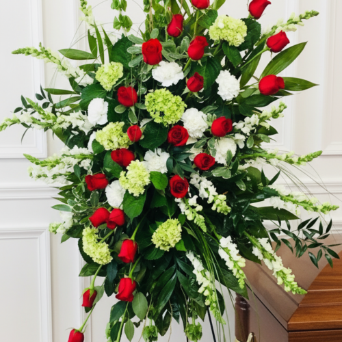 The Forever In Our Hearts Standing Spray, featuring red roses, white blooms, and greenery from The Florist at Plantation, is displayed on a metal easel beside a polished wooden casket in a room with white paneled walls.