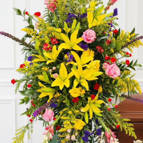 The Led by Light Standing Spray from Clayton Florist showcases yellow lilies, pink roses, red and purple blooms, and greenery on a stand beside a wooden casket in front of a white wall.