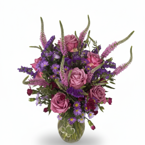 The Enchanted Lavender Garden features a clear glass vase holding a bouquet of purple and lavender roses and asters with green foliage, set against a plain white background.