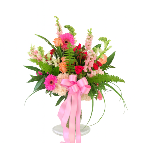 The Peaceful Pink Tribute features pink and peach blooms with green ferns and leaves in a white vase, finished with a large pink ribbon—ideal for Clayton Florist or Clayton NC Flower Delivery.
