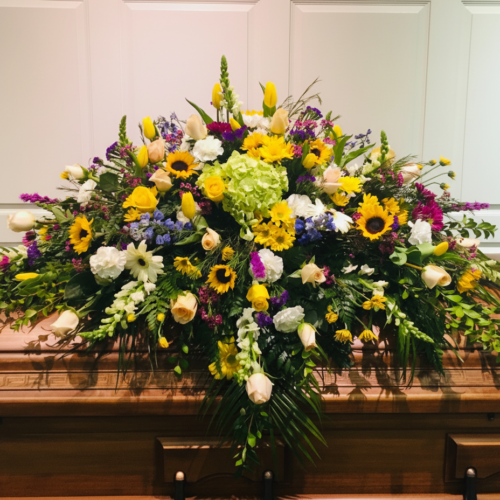 The Warm Horizon Meadows Casket Spray features sunflowers, yellow tulips, white roses, purple blooms, and greenery—artfully arranged by Clayton Florist and displayed on a funeral casket against a white paneled backdrop.