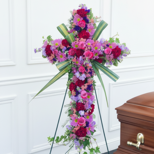 The Everlasting Grace Cross, a large floral cross of pink, purple, and red blooms with lush greenery on a metal easel, stands by a brown wooden casket in a white-walled room—ideal for flower delivery in Clayton, NC.