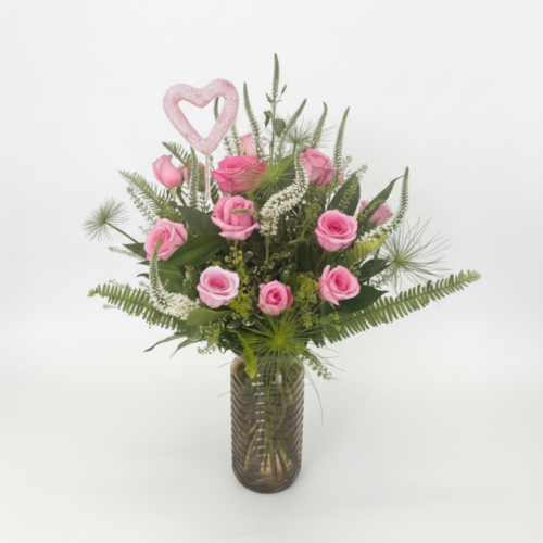 The Head Over Heels Rose Arrangement features a glass vase with pink roses, green ferns, assorted greenery, and a pink heart topper—expertly crafted by your local Clayton florist and available for delivery in Clayton NC.