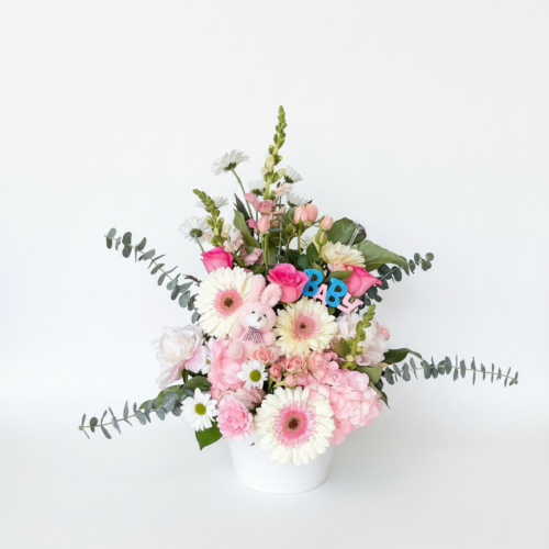 The “It’s A Girl!” arrangement features pink and white flowers, eucalyptus, a plush teddy bear, and blue BABY letters in a white vase—ideal for Clayton NC Flower Delivery from The Florist at Plantation.