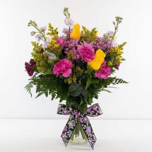 Floral Delight: a glass vase with pink carnations, yellow roses, purple flowers, and greenery, accented by a black floral ribbon—perfect for Flower Delivery in Clayton NC from The Florist at Plantation.