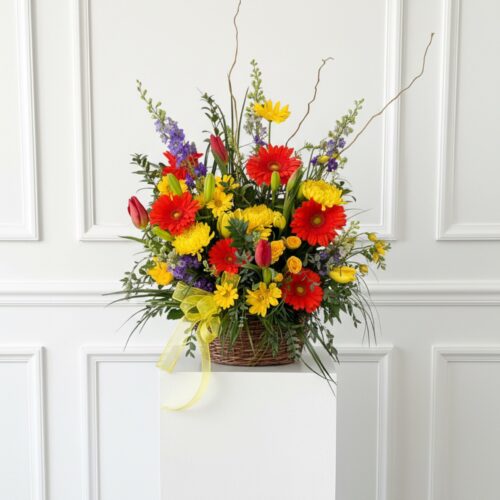 On The Horizon: A vibrant basket of red, yellow, and purple blooms with greenery, elegantly arranged on a white pedestal. Finished with a yellow ribbon by The Florist at Plantation, your trusted local florist in Clayton.