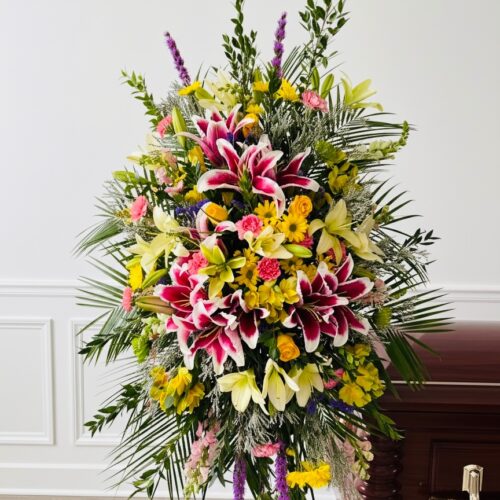 The Luscious Lily Spray by a local Clayton florist, featuring pink lilies, yellow roses, and lush greenery, is displayed beside a closed brown wooden casket in a bright room with white walls and light flooring.