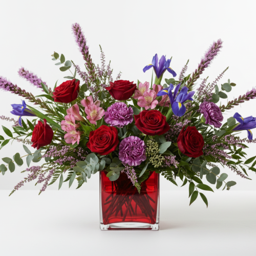 A floral arrangement in a red square vase featuring red roses, purple irises, pink and purple alstroemerias, and sprigs of greenery and lavender, set against a plain white background.