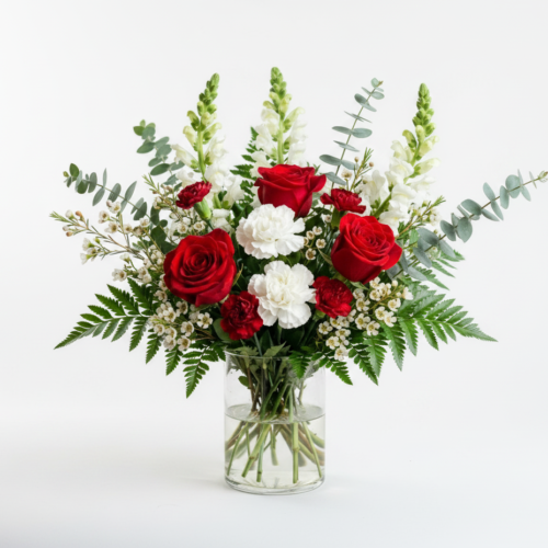 A glass vase filled with red roses, white carnations, small white flowers, snapdragons, ferns, and eucalyptus leaves arranged in a bouquet against a plain white background.