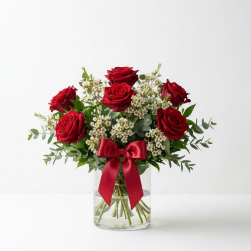 A clear glass vase holds a bouquet of six red roses, white filler flowers, and green foliage, accented with a red satin bow tied around the vase against a plain white background.
